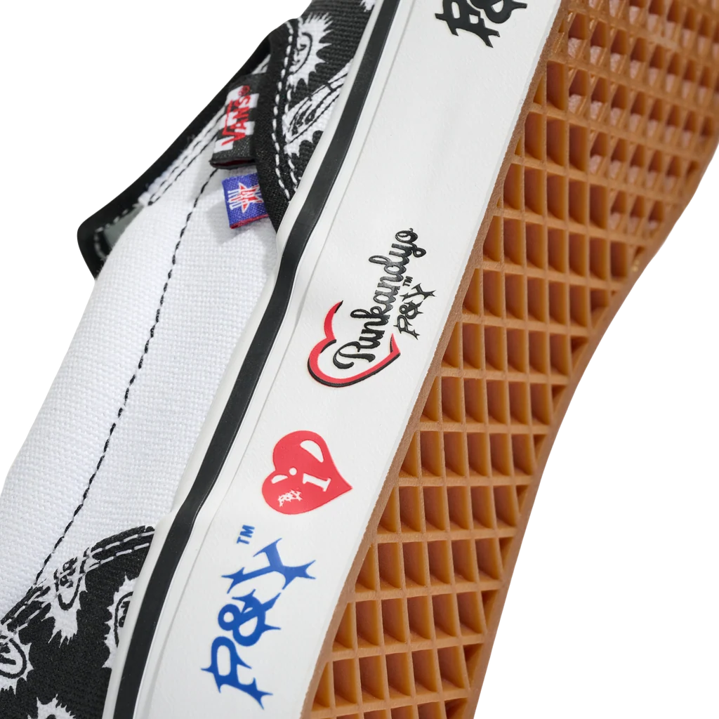 Close-up of a slip-on shoe showing the white canvas upper, black graphic print, white rubber strap with colorful P&H and logos, and a brown waffle sole on a white background.