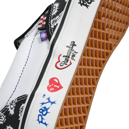 Close-up of a slip-on shoe showing the white canvas upper, black graphic print, white rubber strap with colorful P&H and logos, and a brown waffle sole on a white background.