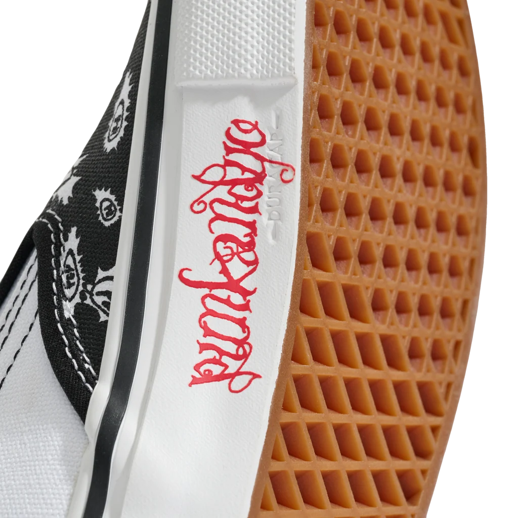 Close-up of a low-top sneaker showing the white midsole with red gothic-style script, a black-and-white patterned canvas side, and a brown gum rubber outsole.