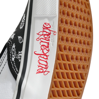 Close-up of a low-top sneaker showing the white midsole with red gothic-style script, a black-and-white patterned canvas side, and a brown gum rubber outsole.