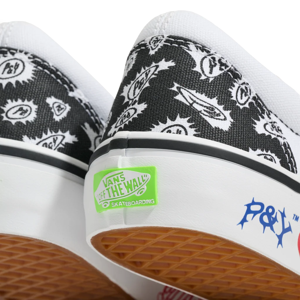 Close-up of the heel area of a pair of Vans 'Off The Wall' skateboarding sneakers, featuring a black and white graphic pattern on the canvas uppers, a white rubber midsole with a green Vans sticker, and a brown textured waffle outsole.