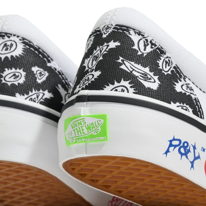 Close-up of the heel area of a pair of Vans 'Off The Wall' skateboarding sneakers, featuring a black and white graphic pattern on the canvas uppers, a white rubber midsole with a green Vans sticker, and a brown textured waffle outsole.