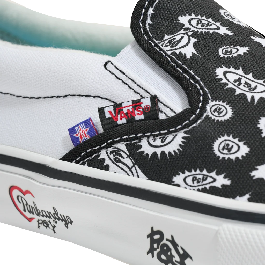 Close-up of a Vans slip-on shoe with a black and white bandana print upper, white rubber sole, and visible Vans and Pink SANDYS collaboration branding.
