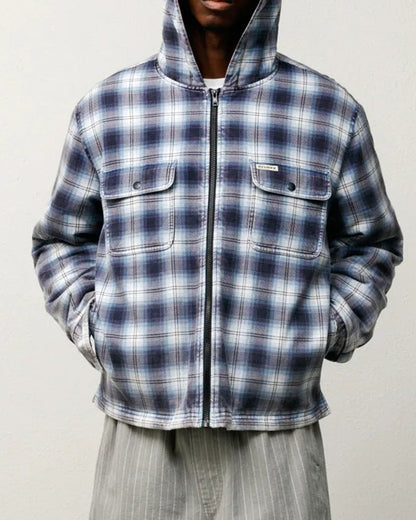 A model stands against a plain off-white wall wearing a blue and white plaid zip-up hooded jacket, light gray pinstriped shorts, white socks, and red sneakers.