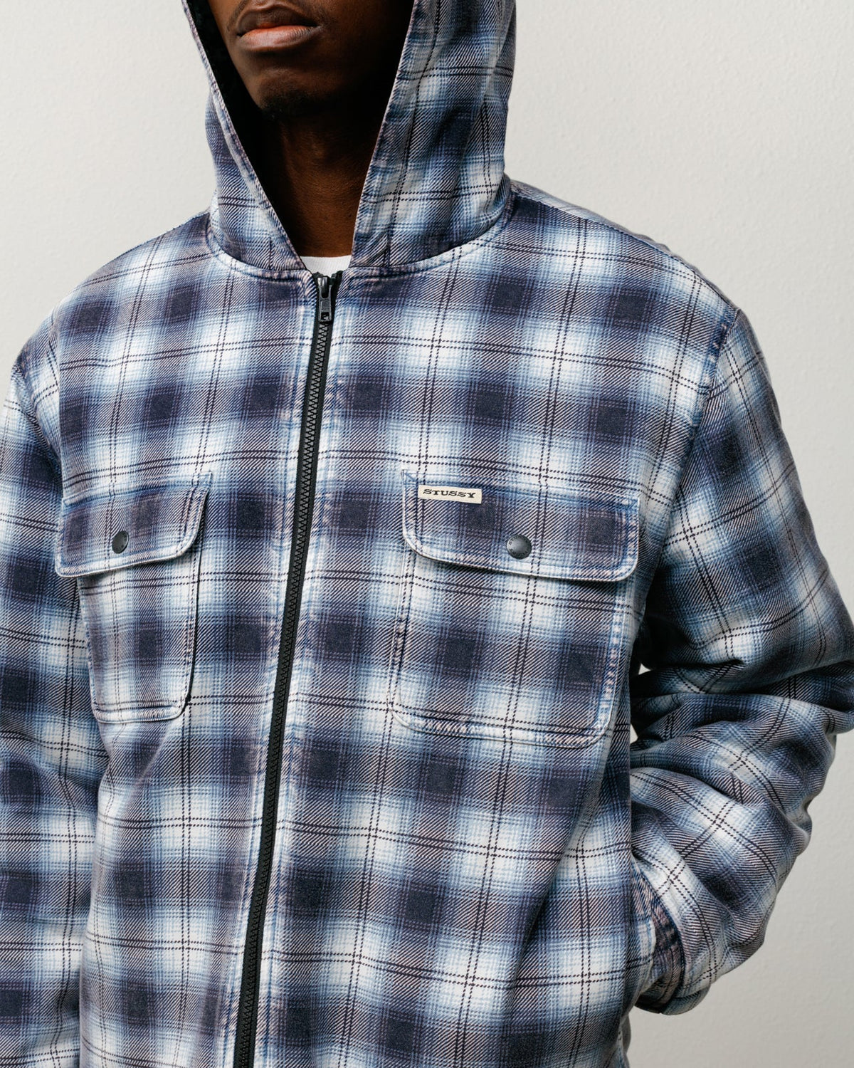 Person wearing a blue and gray plaid jacket with a hood on a light gray background