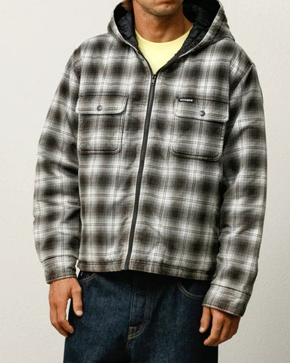 Man wearing a plaid shirt and dark pants standing against a plain background
