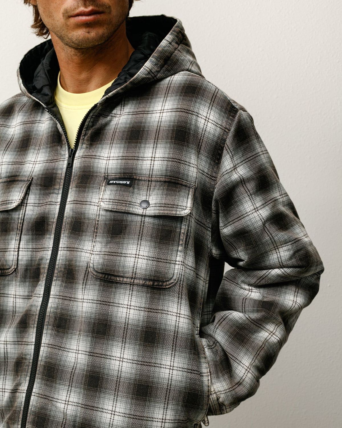 Person wearing a gray and black plaid jacket with a visible brand logo on a plain background