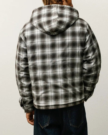 A person viewed from behind wearing a black and white plaid hooded jacket, dark blue jeans, and black shoes, standing against a plain light gray wall and concrete floor.