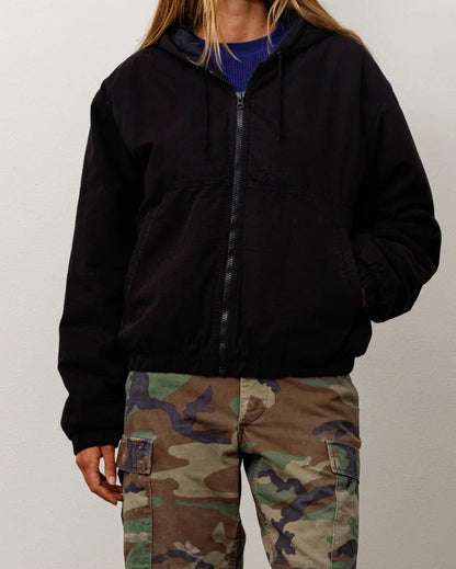 A woman stands against a plain light gray wall wearing a black zip-up hoodie, camouflage cargo shorts, black socks, and hiking boots.