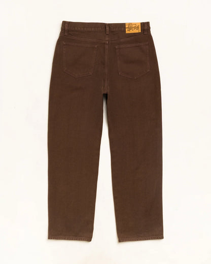 A pair of brown denim jeans shown from the back, featuring two back pockets and a small tan leather brand patch on the waistband, laid flat against a white background.