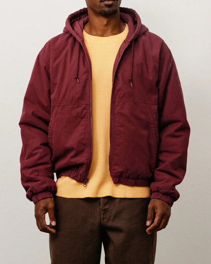 A man stands in a studio wearing a burgundy hooded jacket over a pale yellow shirt, dark brown pants, and brown shoes against a light gray wall and concrete floor.