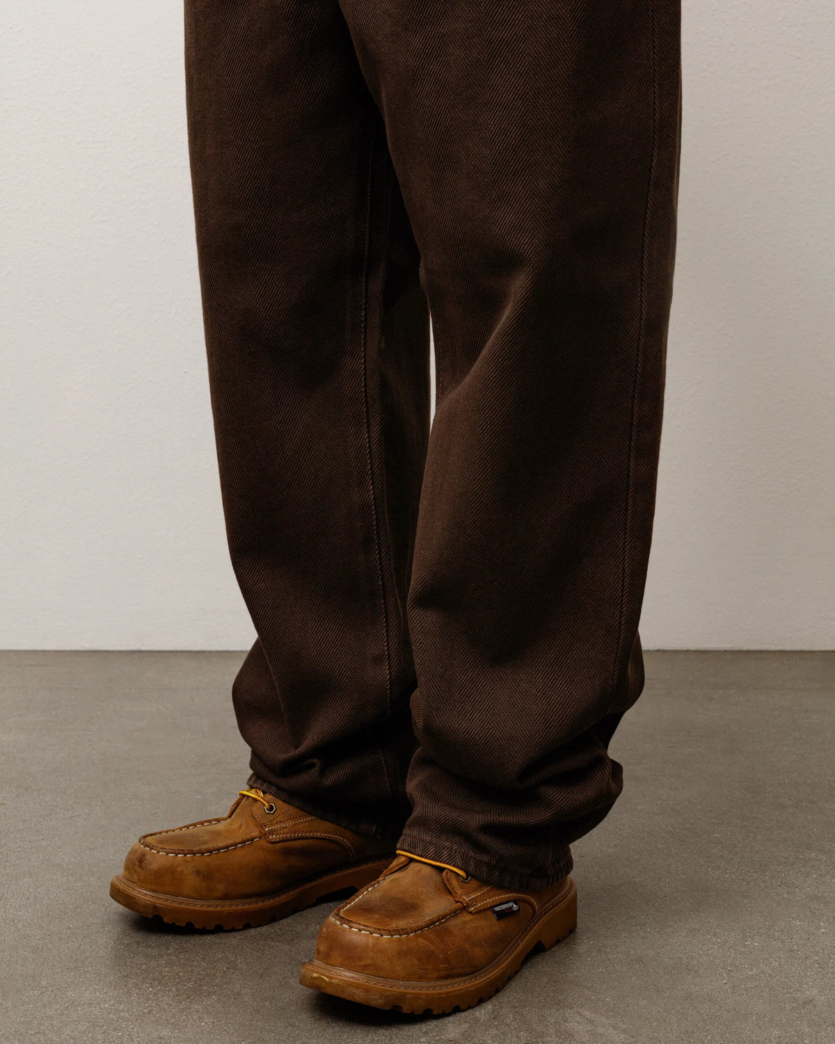 A close-up of a person's legs wearing dark brown work pants and brown suede work boots on a gray concrete floor against a light gray wall.