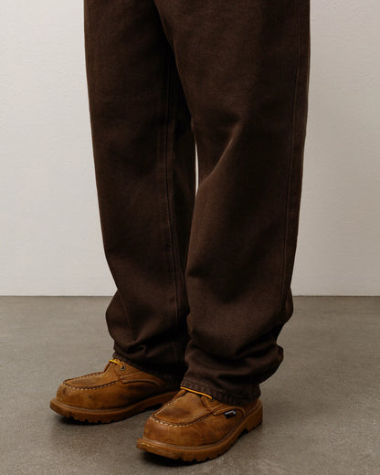 A close-up of a person's legs wearing dark brown work pants and brown suede work boots on a gray concrete floor against a light gray wall.