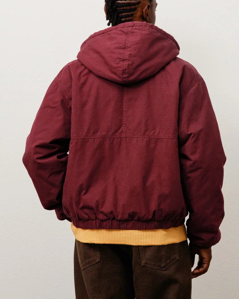 A person viewed from behind wearing a maroon hooded puffer jacket over a yellow shirt, dark brown pants, and brown shoes, standing against a plain light gray wall.