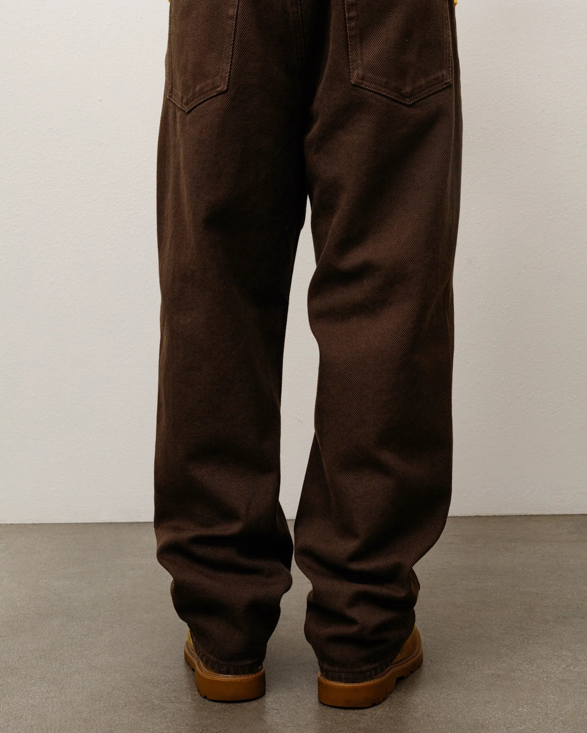 Rear view of a person wearing dark brown wide-leg pants and brown shoes against a plain light gray wall and concrete floor.
