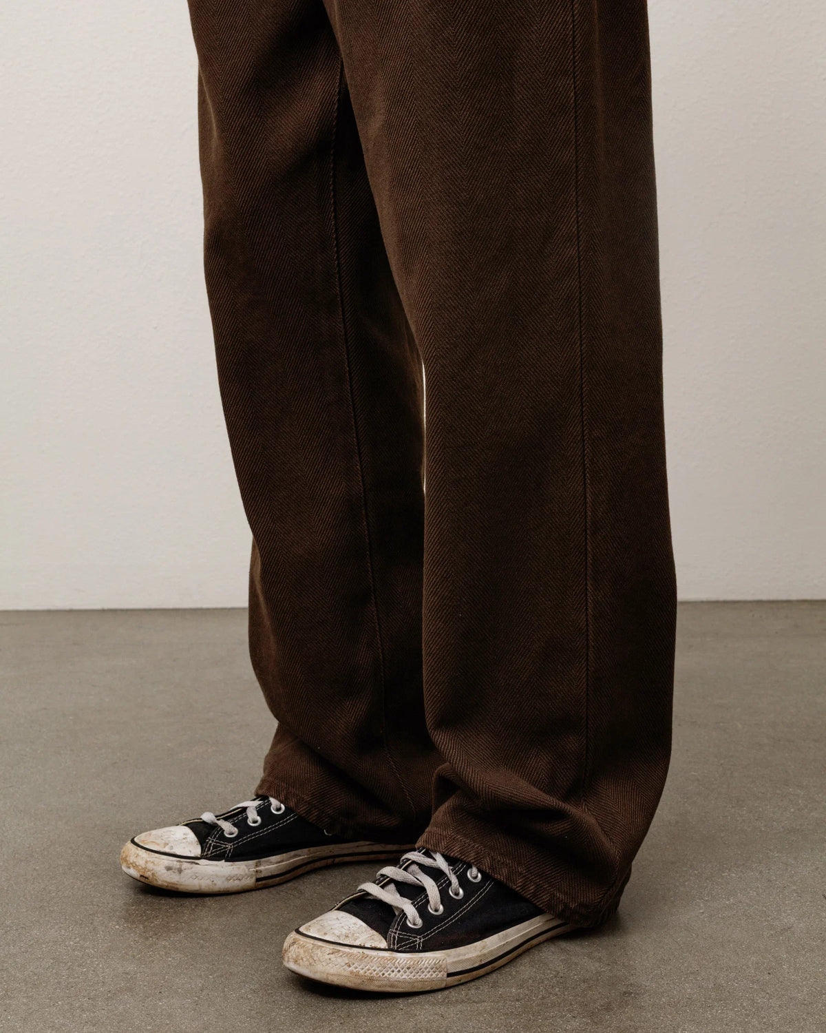 A close-up of a person's legs wearing dark brown wide-leg trousers and black canvas low-top sneakers with white rubber toe caps, standing on a gray concrete floor against a light gray wall.