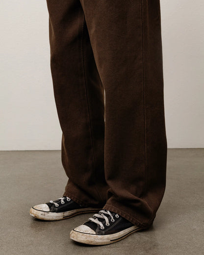 A close-up of a person's legs wearing dark brown wide-leg trousers and black canvas low-top sneakers with white rubber toe caps, standing on a gray concrete floor against a light gray wall.