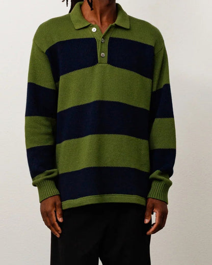A man wearing a green and navy blue horizontally striped long-sleeve polo sweater, black trousers, and black sandals, standing against a plain off-white wall.