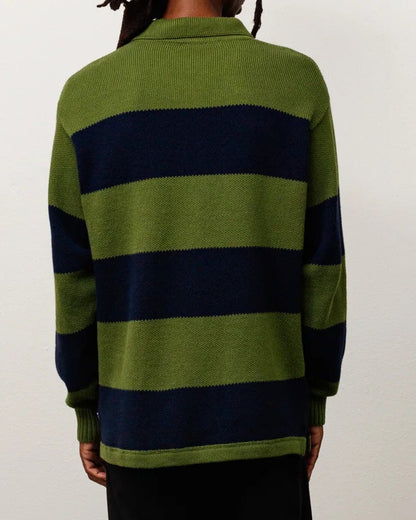 A man viewed from behind wearing a green and navy blue horizontally striped knit sweater, black trousers, and black sandals, standing against a plain off-white wall.