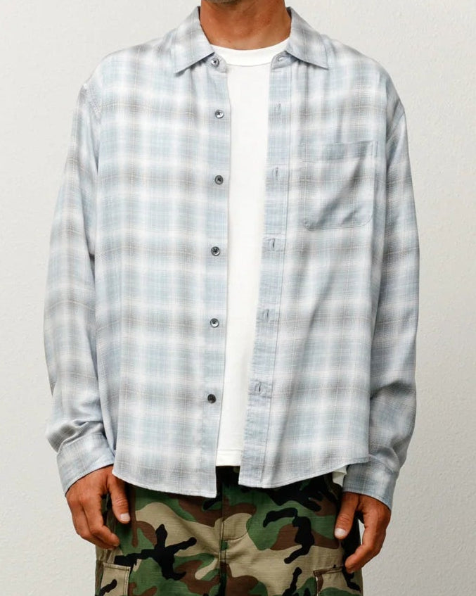 A man stands against a plain light gray wall wearing a light blue and white plaid button-up shirt over a white t-shirt, green camouflage cargo pants, and a beige baseball cap.