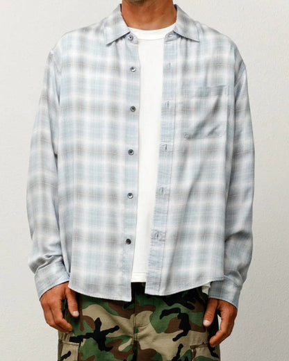 A man stands against a plain light gray wall wearing a light blue and white plaid button-up shirt over a white t-shirt, green camouflage cargo pants, and a beige baseball cap.