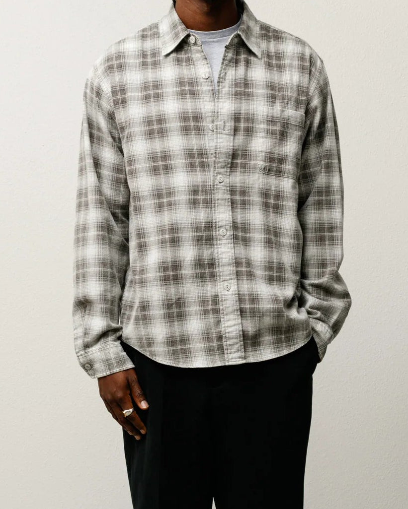 A man stands against a plain off-white wall wearing a long-sleeve plaid button-up shirt in gray and white, black trousers, and black shoes.