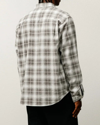 Person wearing a plaid shirt and dark pants standing against a white wall.