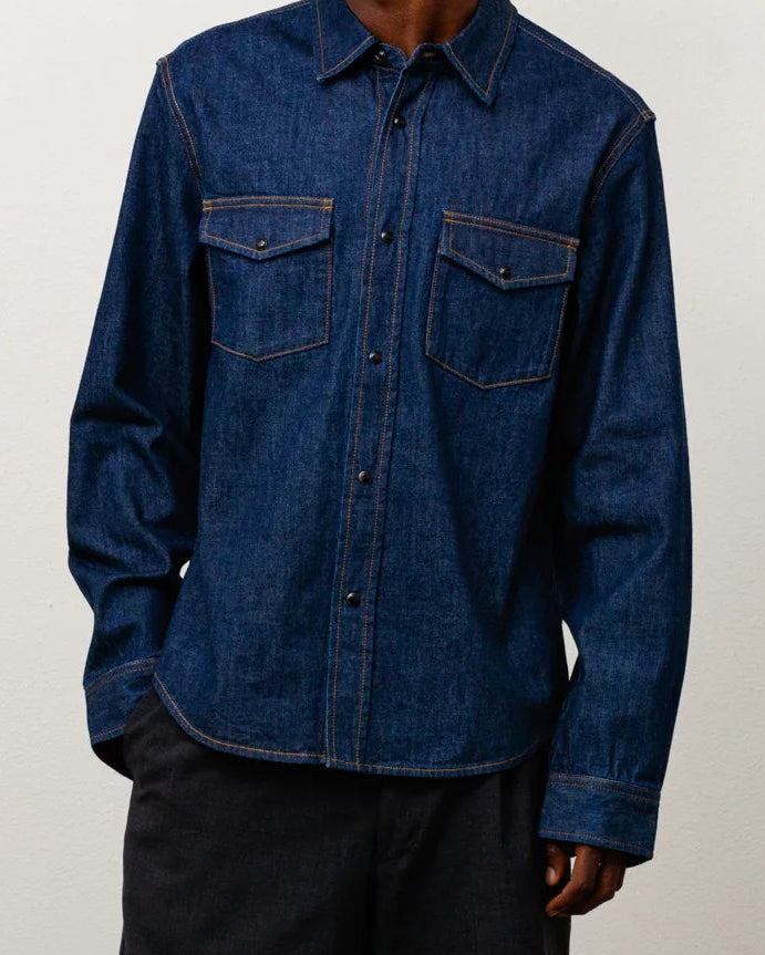 A model stands in a studio wearing a dark blue denim button-up shirt, dark charcoal shorts, white socks, and brown boots against a plain off-white wall and concrete floor.