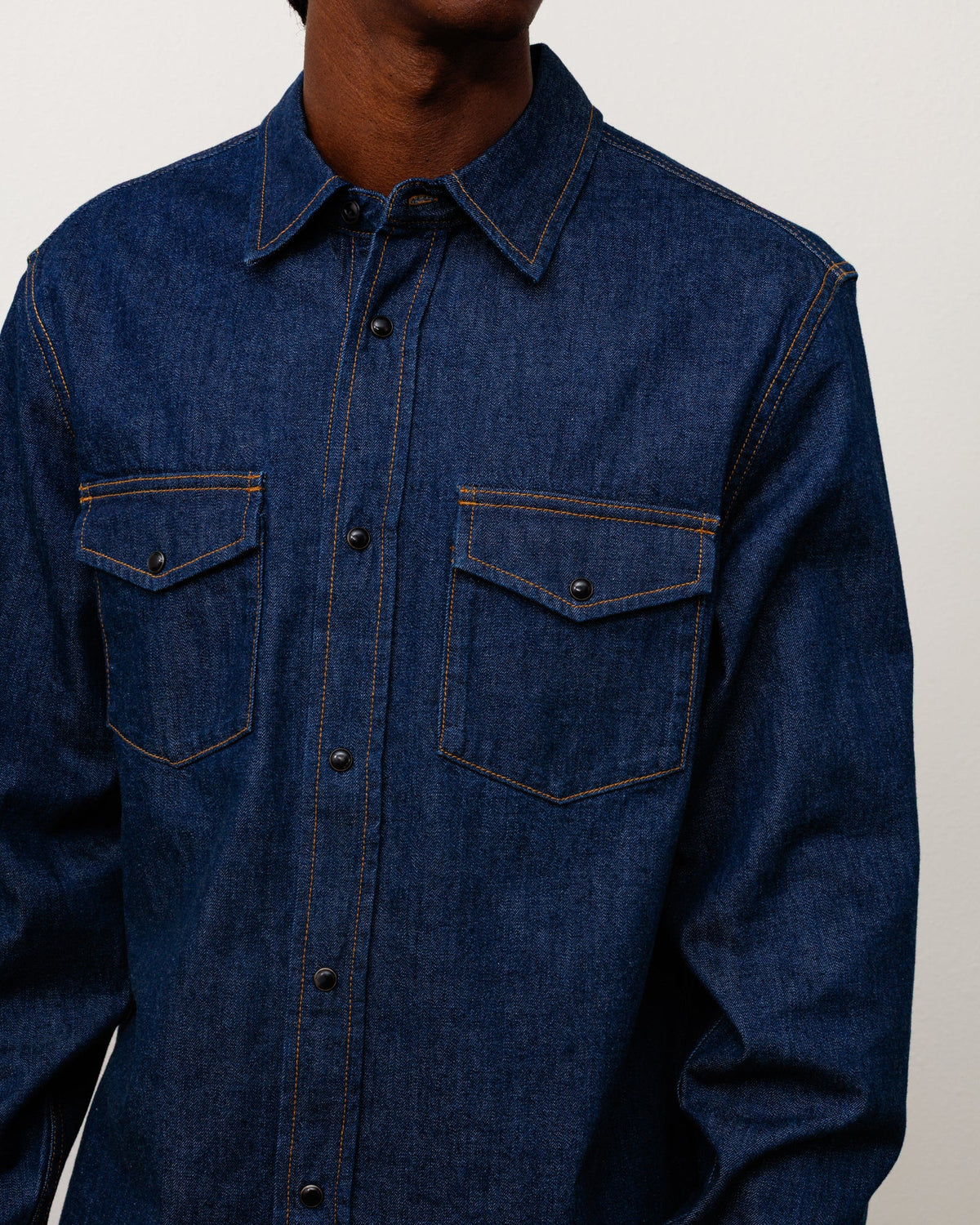 A close-up of a person wearing a dark blue denim button-up shirt with two chest pockets and contrast orange-brown stitching, against a plain light background.