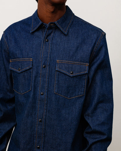 A close-up of a person wearing a dark blue denim button-up shirt with two chest pockets and contrast orange-brown stitching, against a plain light background.