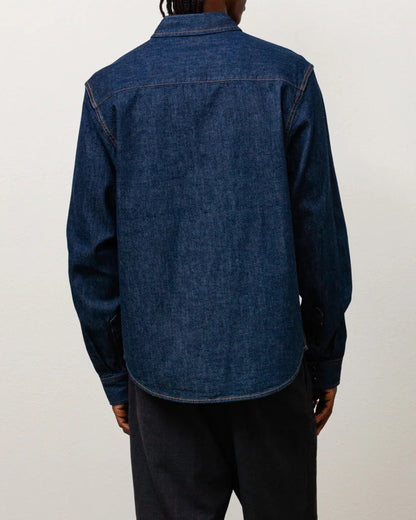 A model viewed from behind wearing a dark blue denim long-sleeve button-up shirt, dark charcoal shorts, white socks, and brown boots, standing against a plain off-white wall and concrete floor.