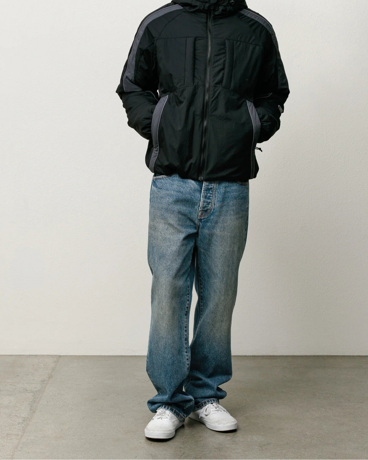 A man wearing a black hooded jacket with gray side panels, blue jeans, and white sneakers, standing against a plain light gray studio wall.