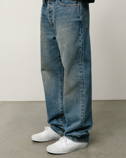 A person wearing loose-fit medium-wash blue denim jeans with white low-top sneakers, shown from the waist down against a plain light gray wall and concrete floor.
