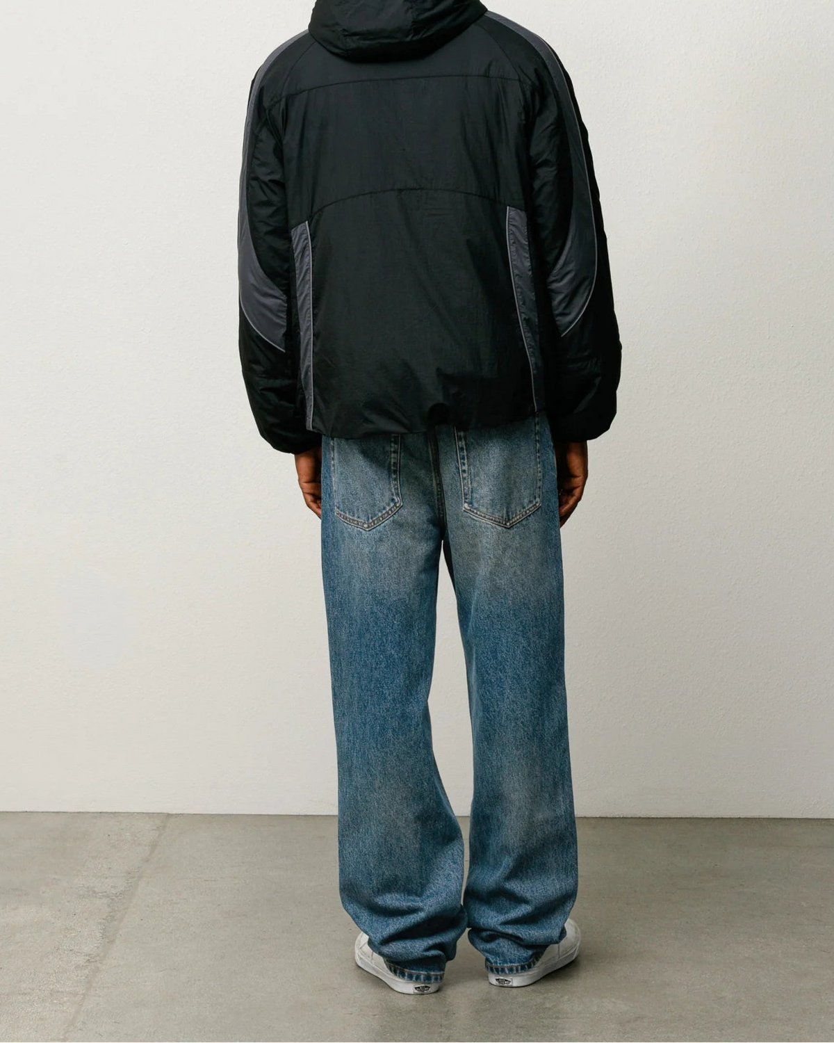 A person shown from the back wearing a black hooded jacket with dark gray side panels, blue jeans, and white sneakers, standing against a plain off-white wall.
