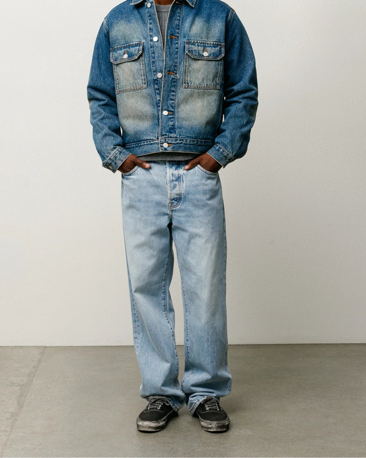 A man modeling a blue denim jacket and light-wash denim jeans against a plain light gray studio background.