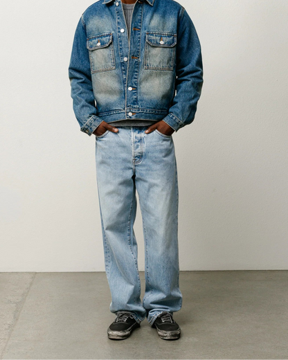 A man modeling a blue denim jacket and light-wash denim jeans against a plain light gray studio background.