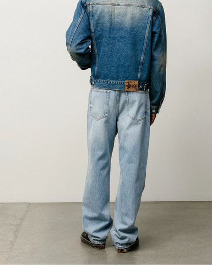 A model viewed from behind wearing a blue denim jacket and light-wash blue jeans, standing against a plain off-white wall and concrete floor.