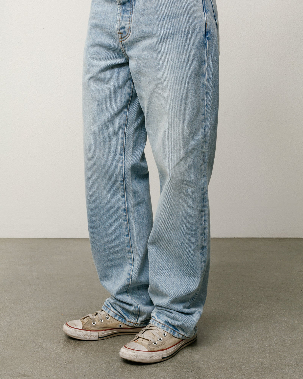 Light-wash blue denim jeans worn by a model, paired with beige low-top sneakers, shown from the waist down against a neutral studio background.