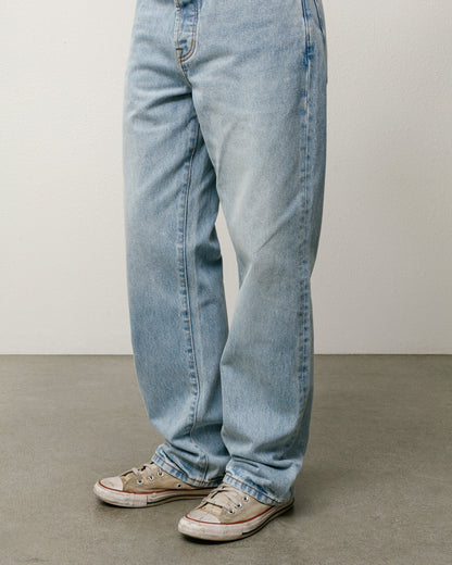 Light-wash blue denim jeans worn by a model, paired with beige low-top sneakers, shown from the waist down against a neutral studio background.