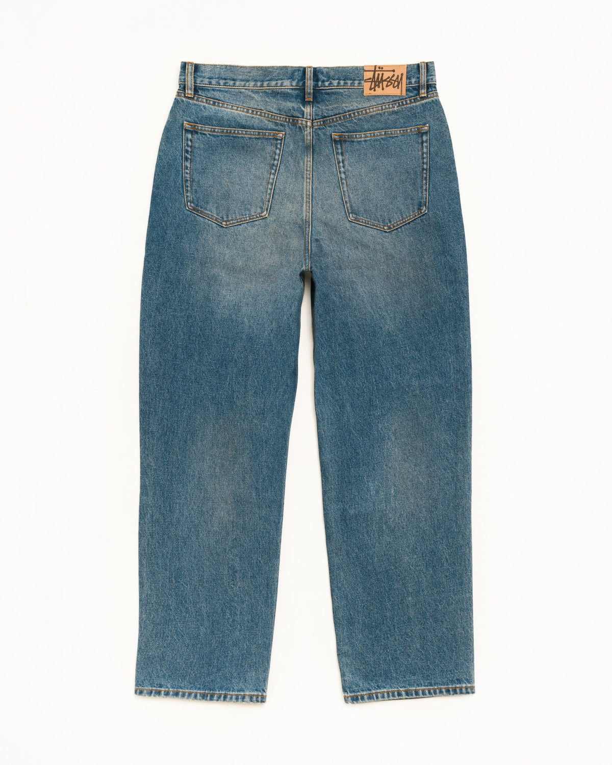Back view of a pair of blue denim jeans with a straight-leg fit, featuring classic five-pocket styling and a visible Stüssy brand patch on the back waistband.