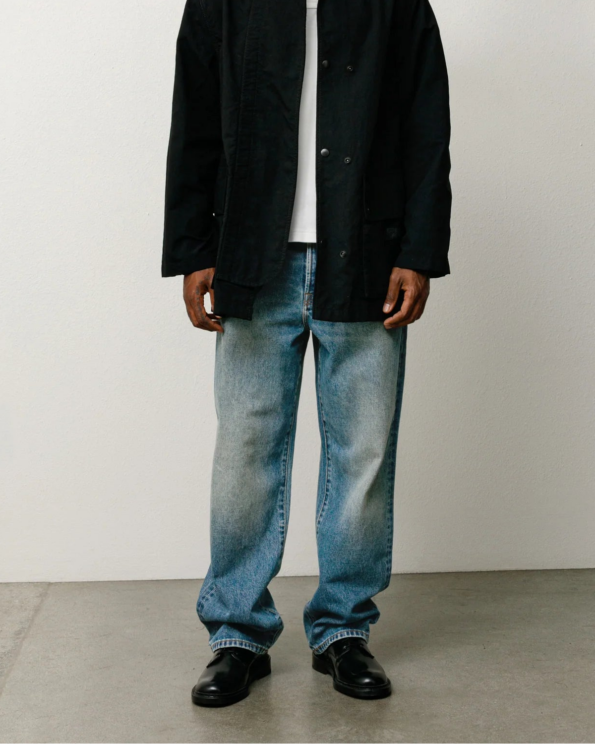 A man stands against a plain light gray wall wearing a black overshirt over a white collared shirt, light blue faded jeans, and black shoes.