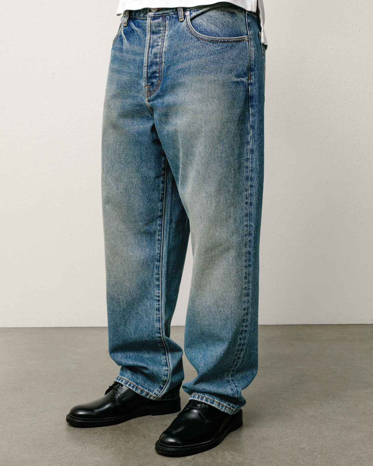 A person wearing loose-fit medium-wash blue denim jeans with black leather shoes, shown from the waist down against a plain light gray wall and concrete floor.