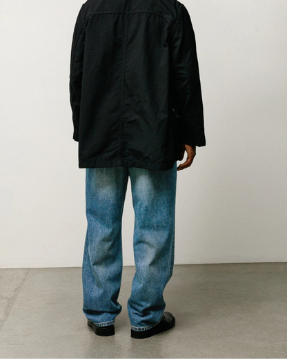 A man viewed from behind wearing a black overshirt, blue jeans, and black shoes, standing against a plain off-white wall and concrete floor.
