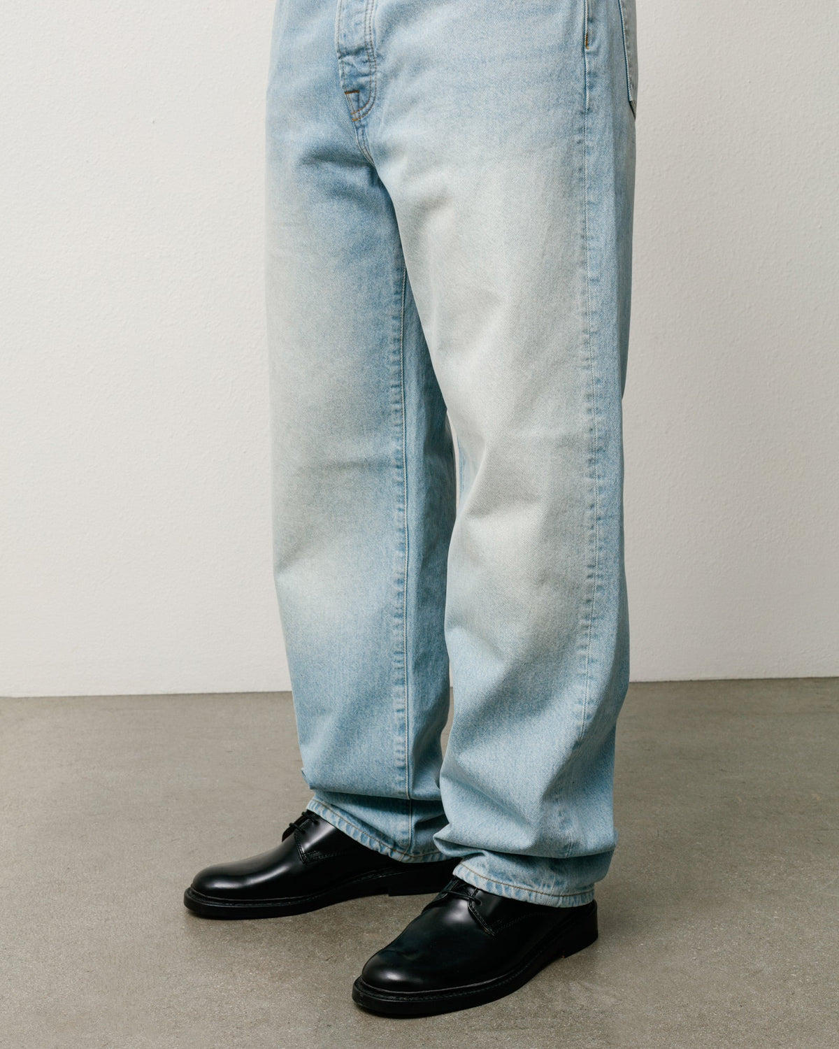 Light wash blue denim jeans shown from the knees down, worn with black leather shoes on a neutral studio floor and wall.