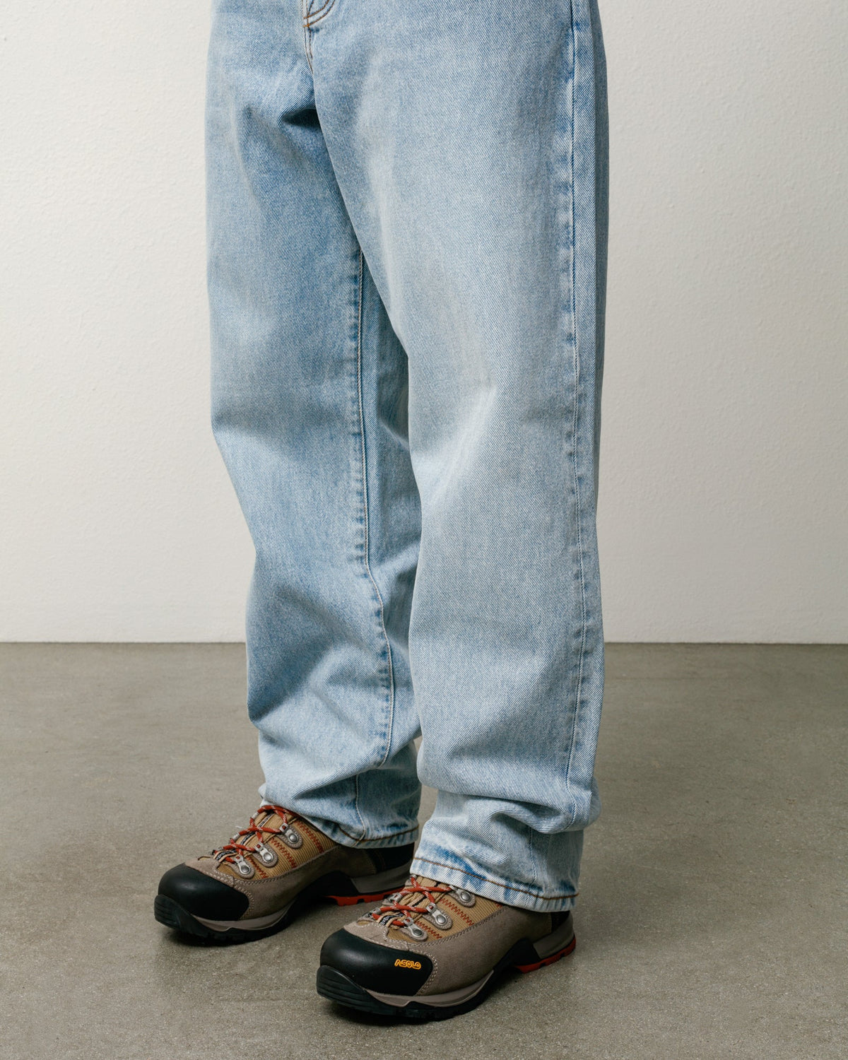 Close-up of a person's legs wearing light-wash blue jeans and brown hiking boots with black toes, standing on a gray concrete floor against a plain off-white wall.