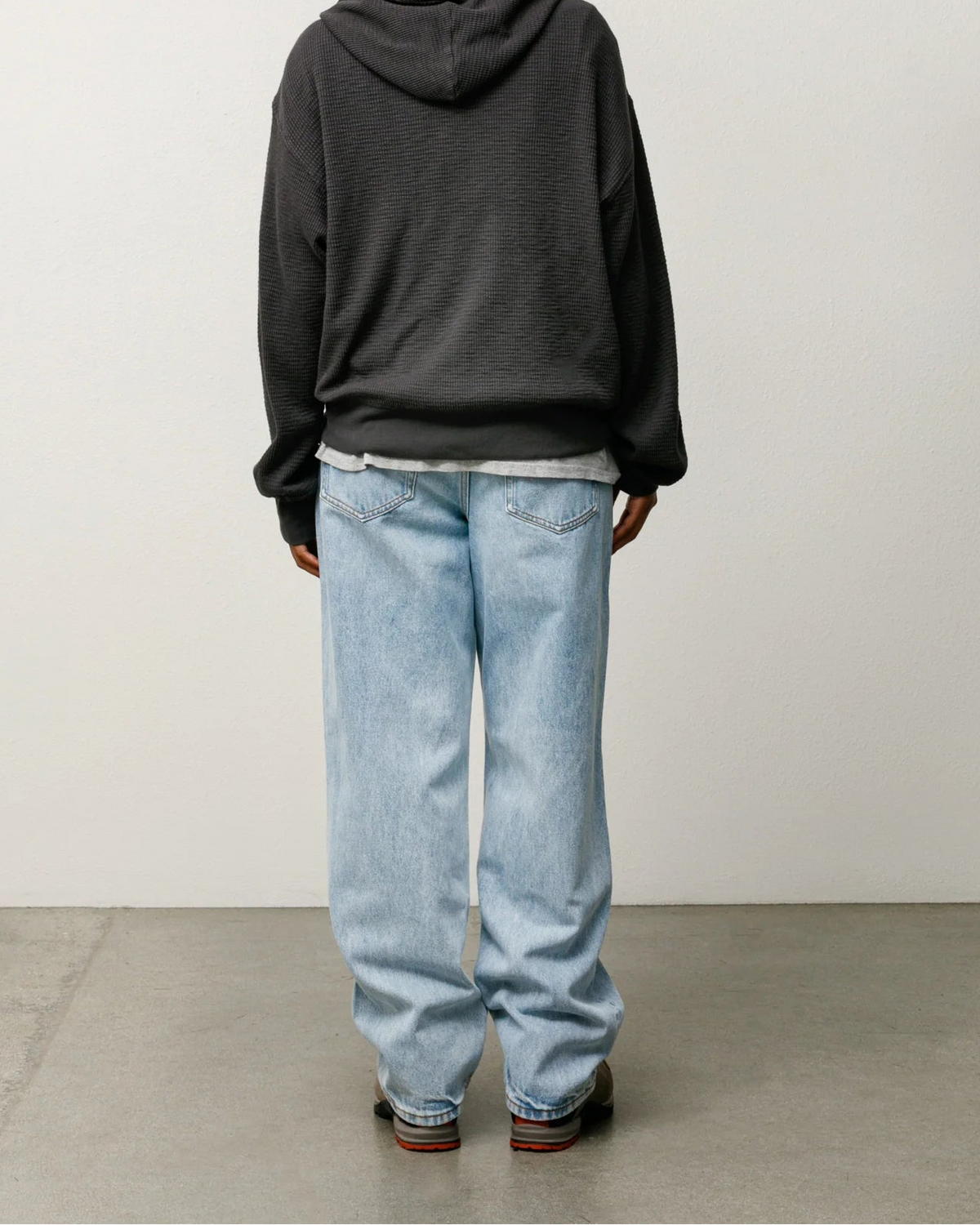 A person shown from the back wearing an oversized dark charcoal gray hoodie, light-wash blue jeans, and brown shoes, standing against a plain light gray wall and concrete floor.