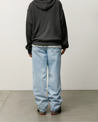 A person shown from the back wearing an oversized dark charcoal gray hoodie, light-wash blue jeans, and brown shoes, standing against a plain light gray wall and concrete floor.