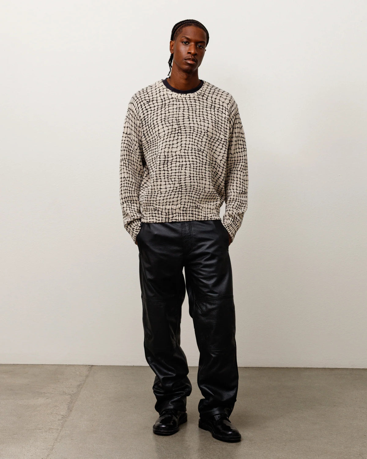 A model stands in a studio wearing a beige and black textured knit sweater, black leather pants, and black shoes against a plain off-white wall and concrete floor.