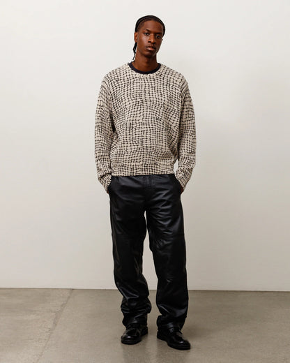 A model stands in a studio wearing a beige and black textured knit sweater, black leather pants, and black shoes against a plain off-white wall and concrete floor.