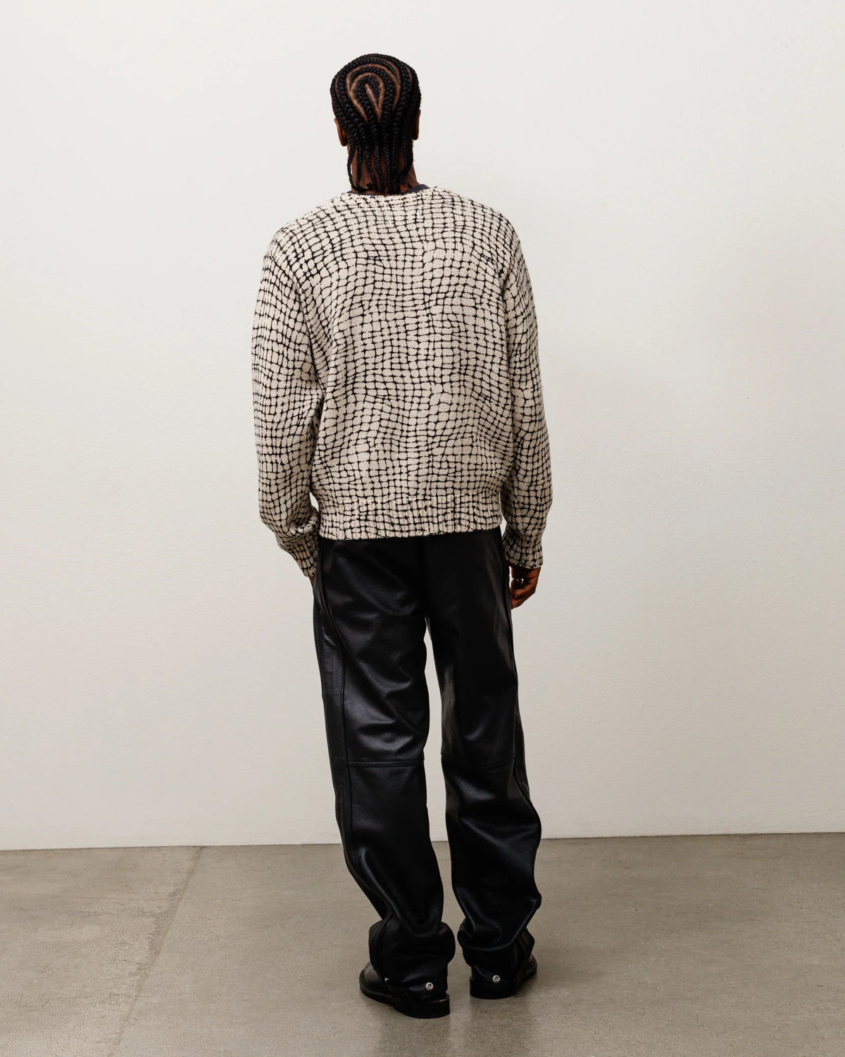 A model viewed from behind wearing a beige and black patterned knit sweater and black leather pants, standing against a plain off-white wall on a concrete floor.